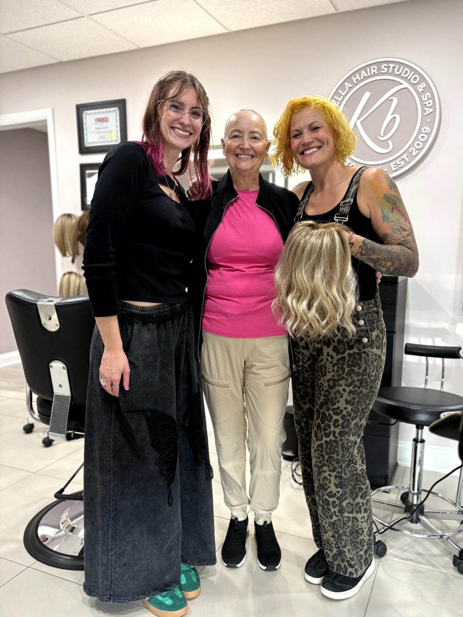 Client wearing a blonde wavy wig stands between two women in the K Bella Hair Studio & Spa salon in Brighton, MI, by Kristan Sayers at K Bella Hair Studio & Spa in Brighton, MI