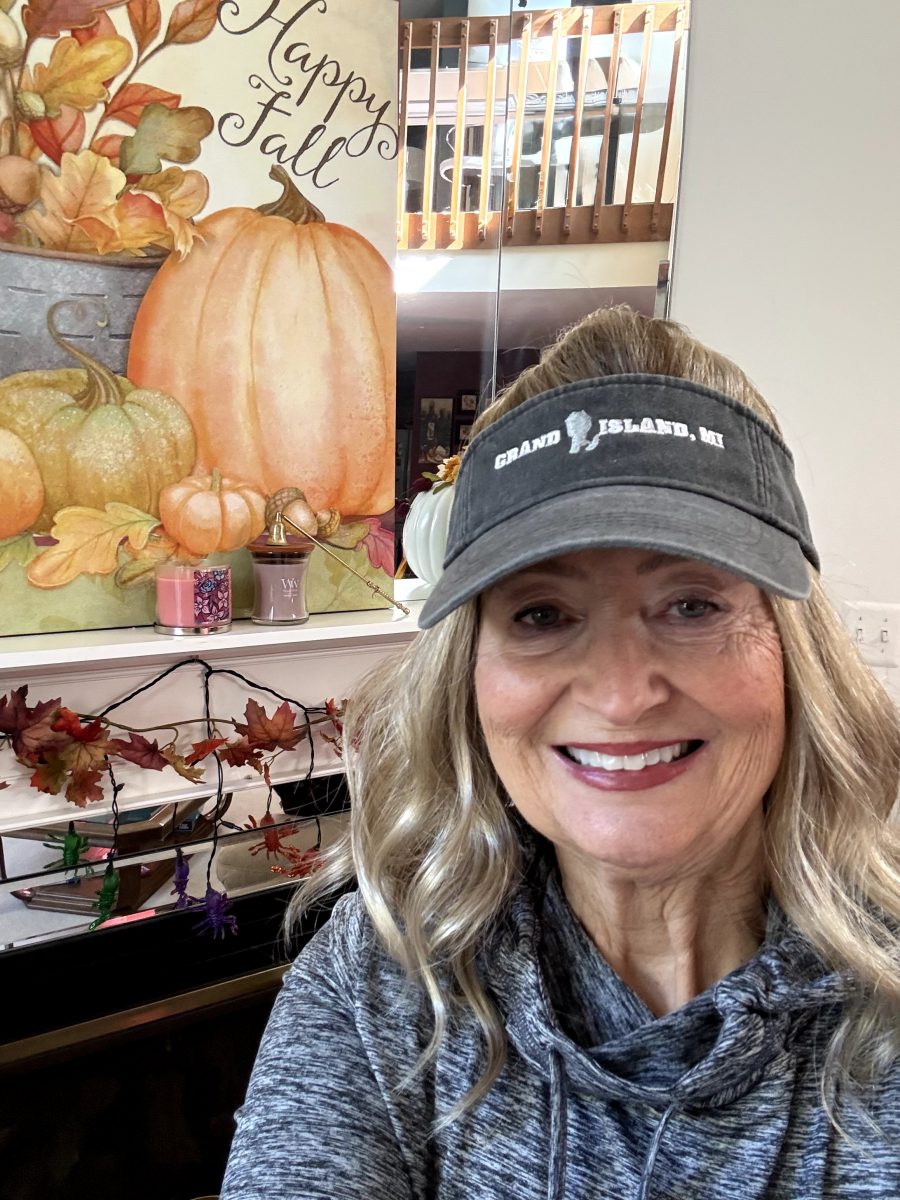 A woman wearing a blonde wavy wig and black visor, smiling at the camera in the salon, with autumn decorations visible in the background, by Kristan Sayers at K Bella Hair Studio & Spa in Brighton, MI