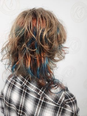 Back view of shoulder-length layered hair with vivid blue and red streaks and thin rattails, worn over plaid shirt, by Siobhan Rozinski at K Bella Hair Studio & Spa in Brighton, MI