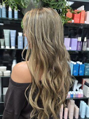 Long wavy dark blonde to brunette hair with fusion extensions, shown from behind, by Kristan Sayers at K Bella Hair Studio & Spa in Brighton, MI