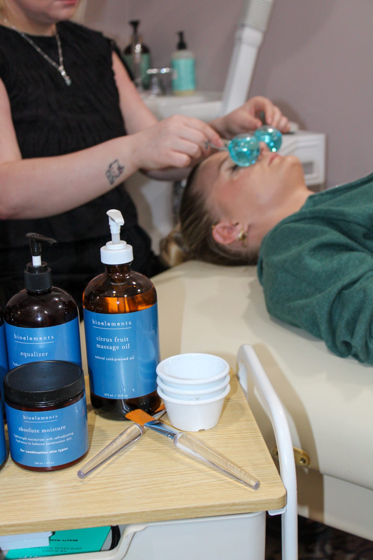 Professional Bioelements cleanser, massage oil and moisturizer on the treatment cart beside a facial in progress at K Bella Hair Studio in Brighton, Michigan